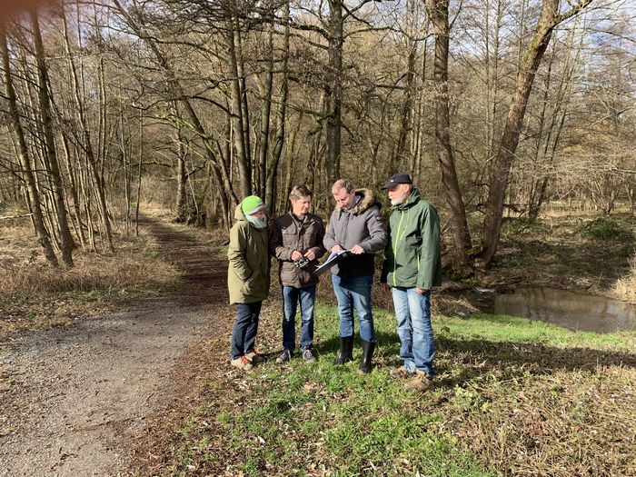 Vier Personen stehen neben einem Weiher und betrachten einen Plan.