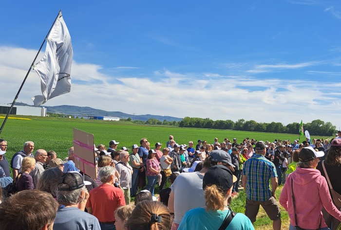 Viele Demonstrierende stehen an einer Wiese, im Hintergrund ein Gewerbegebiet.